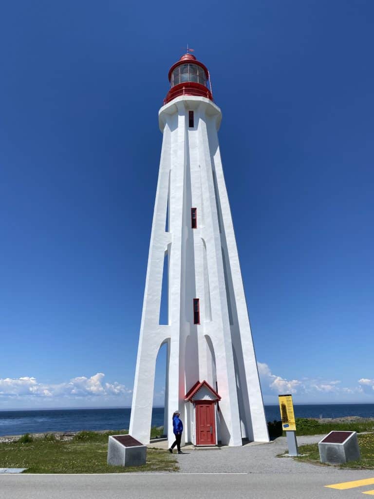 Woman walking in front of Point-au-Pere lighthouse which is white with red light and red door in Rimouski, Quebec with St. Lawrence in background.