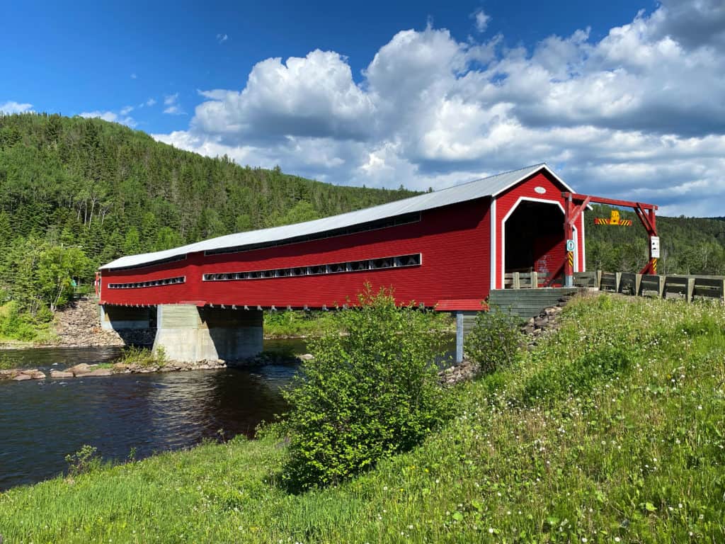 Bright red wooden covered bridge over river with blue sky and fluffy white clouds in Routhierville, Quebec on Gaspe road trip.