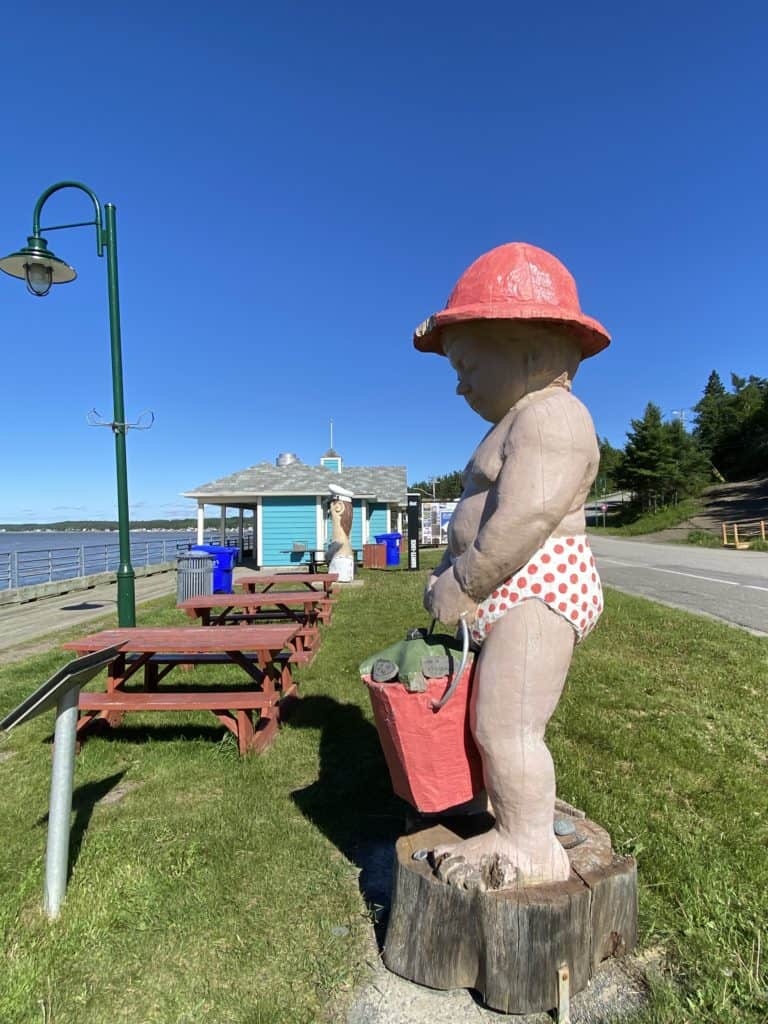 large sculpture of toddler in red hat, red polka-dot bathing suit bottoms carrying red pail along boardwalk in Sainte-Luce, Quebec with light, row of picnic tables and bright blue buildings