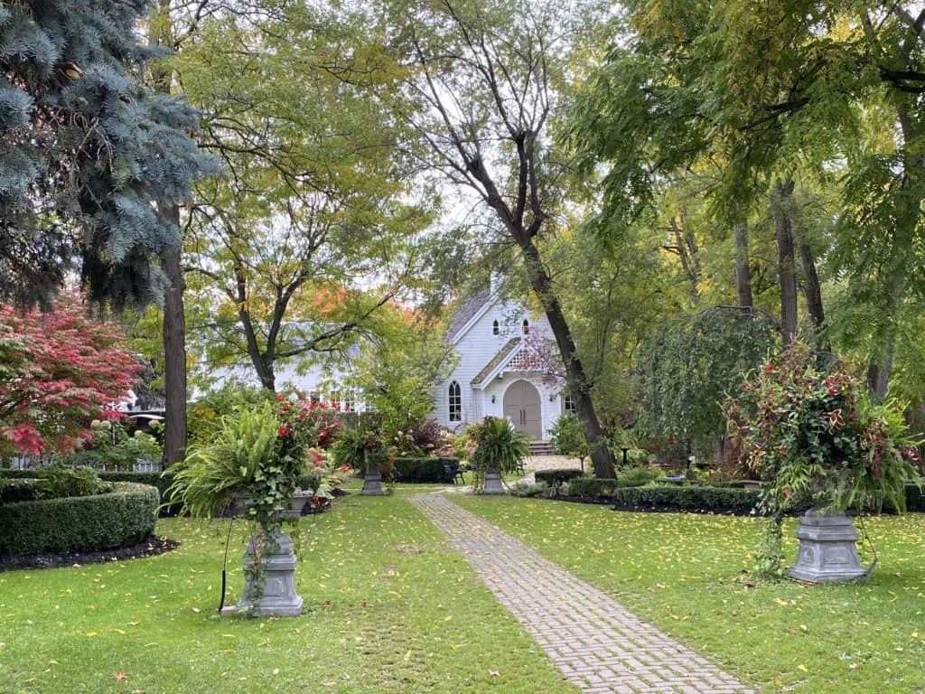 Garden in Kleinburg with white church in background partly hidden by trees and bushes.