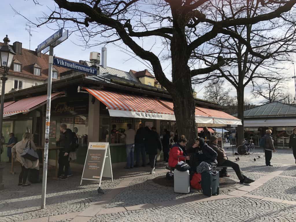 Street sign reading "Viktualienmarkt" in front of market stall in Munich, Germany. People walking on street and sitting by tree with luggage.