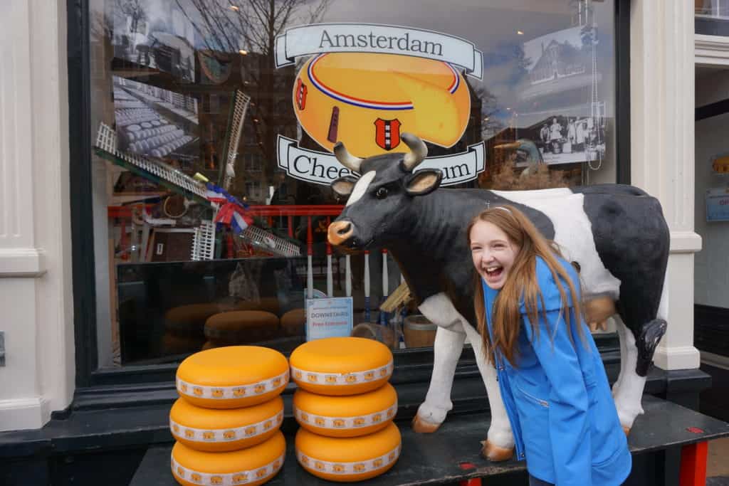 Young girl in blue coat laughing at display with cow and 6 stacked rounds of cheese outside the Amsterdam Cheese Museum.