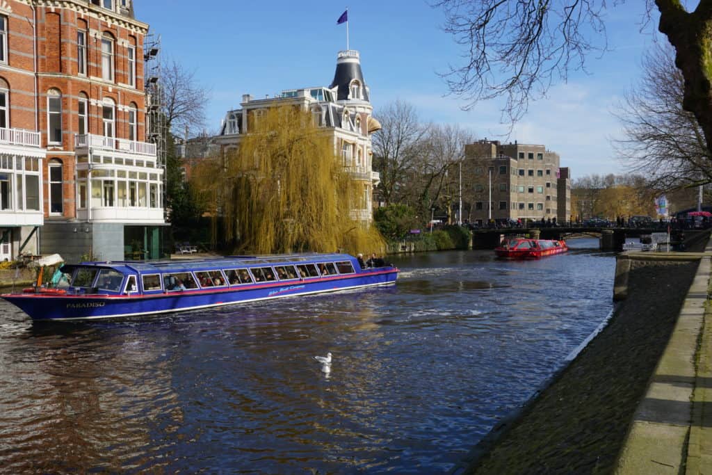 Blue canal boat and red canal boat behind cruising on a canal in Amsterdam.