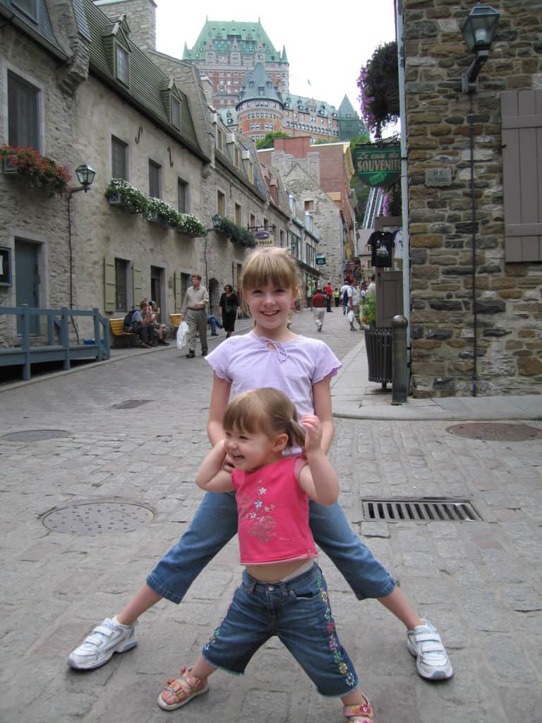 Two young girls standing on a street in the lower town of Quebec City with stone buildings and Chateau Frontenac in the background.