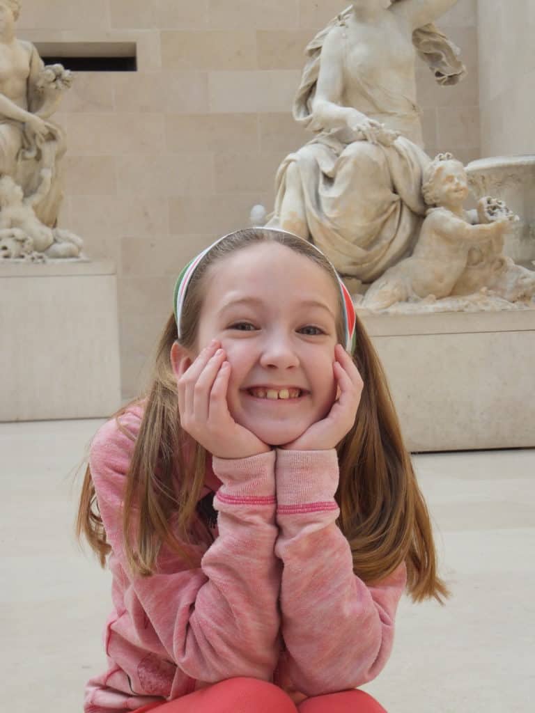 Young girl in pink with hands on face sitting in a courtyard at the Louvre museum with white marble statues in background.