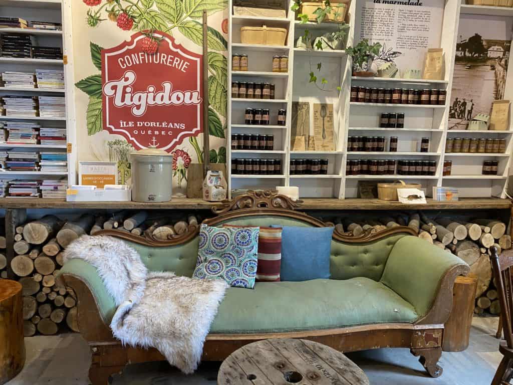 Tigidou shop on Ile d'Orleans, Quebec - green settee with cushions in front of stacked fiewood and white shelves with books, jars of jam and other items.