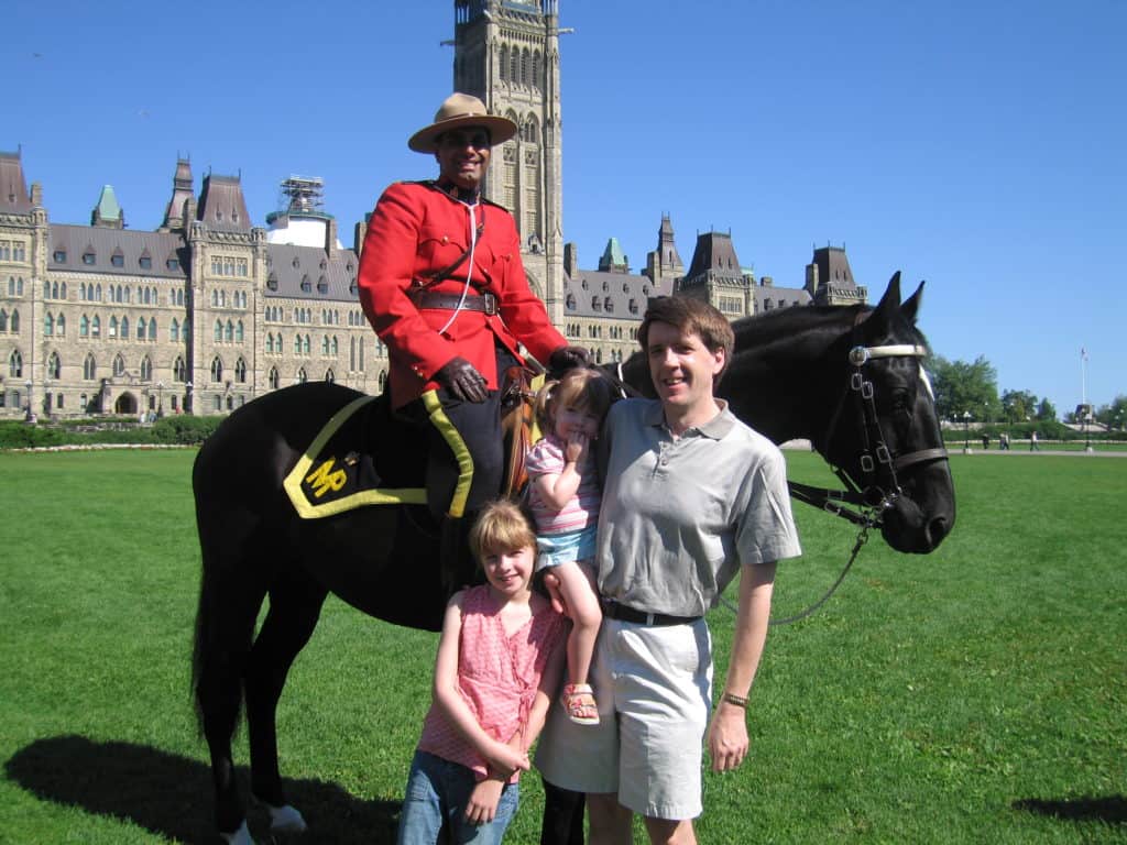 Young girl and man holding toddler girl standing with Royal Canadian Mounted Police officer on horseback in front of Canadian Parliament building in Ottawa.