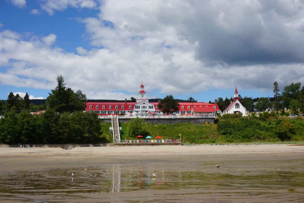 View of Tadoussac, Quebec from water - beach with stairs up to red and white historic Tadoussac hotel and small church.
