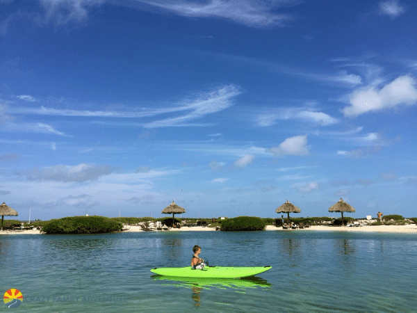 Person in bright green kayak in water at Hawks Cay Resort with beach and umbrellas in background.