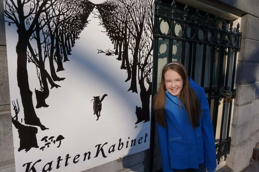 Young girl in blue coat in front of black and white sign for KattenKabinet museum in Amsterdam.