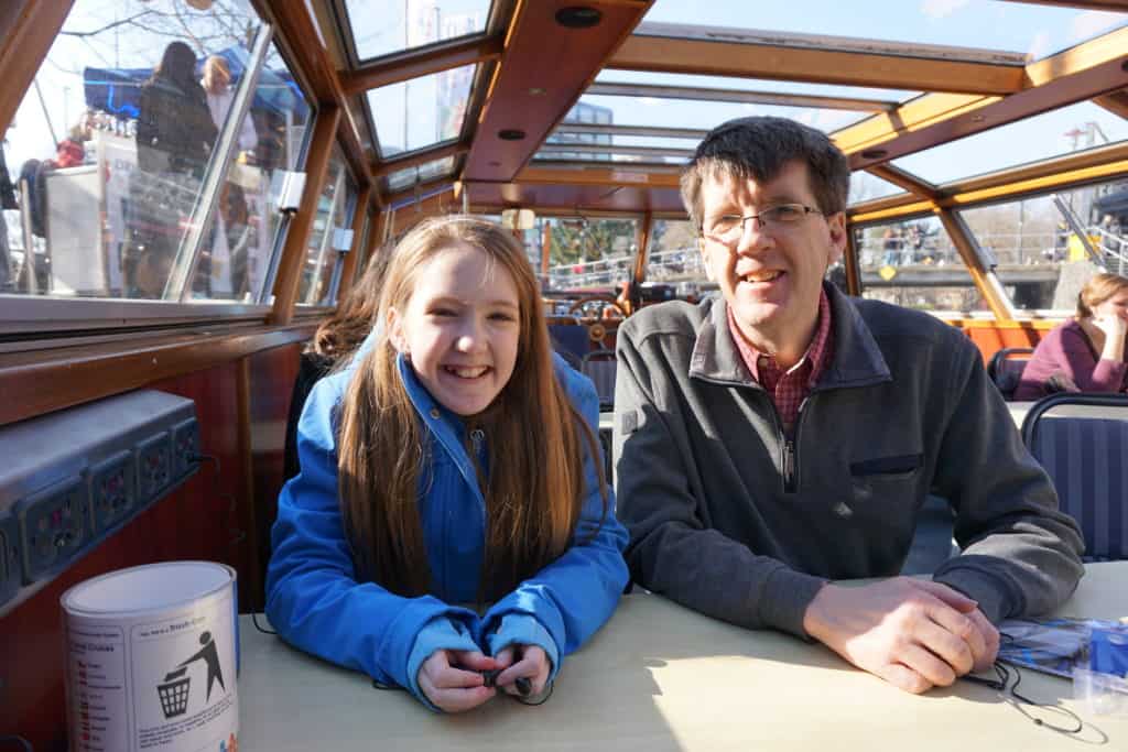 Young girl in blue coat and man wearing glasses and grey sweater sitting at a table in a canal boat with glass roof in Amsterdam.