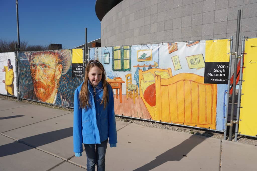 Young girl in blue coat in front of a fence with images of Van Gogh paintings outside the Van Gogh Museum in Amsterdam.