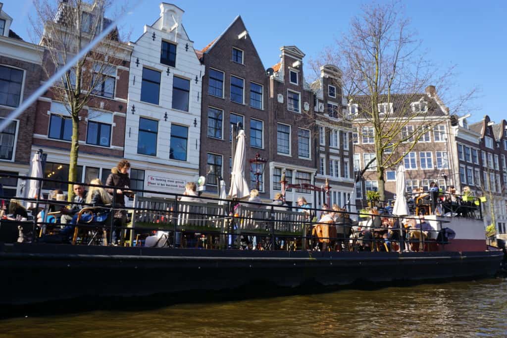 View from a canal cruise in Amsterdam - canal houses and people sitting out at tables alongside the canal.