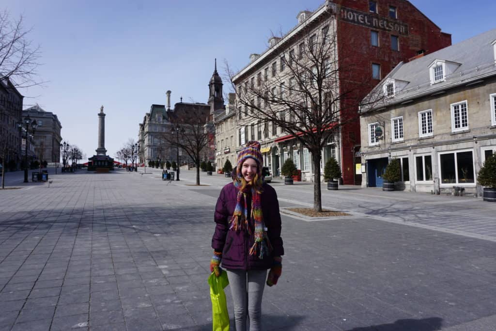 Young girl in purple winter coat, multicoloured hat and mittens holding green shopping bag in Place Jacques Cartier in Old Montreal.