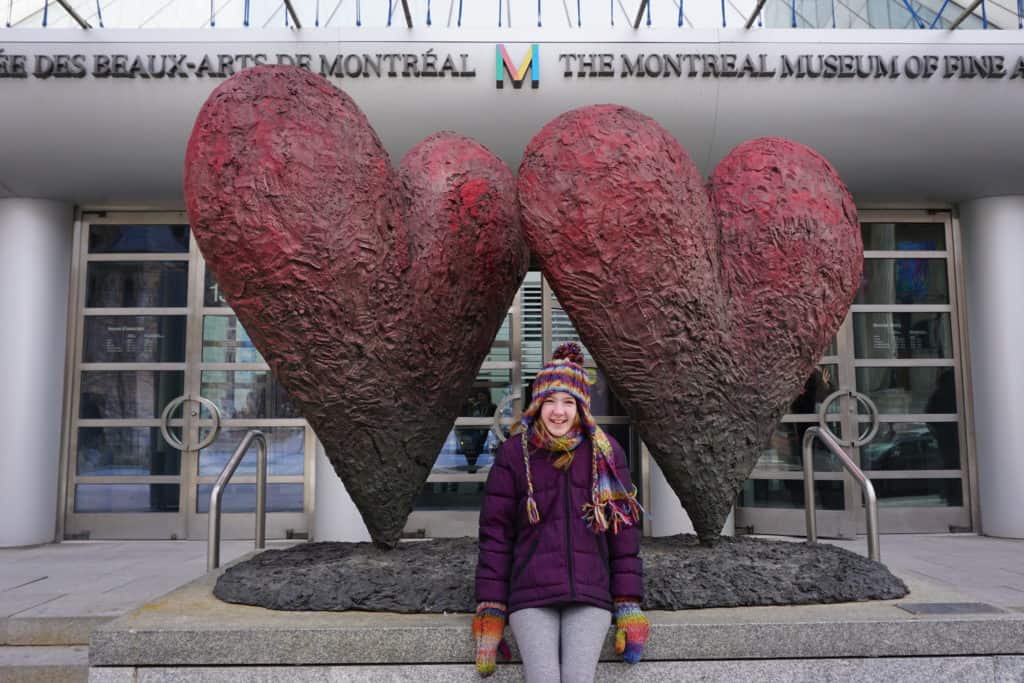 Young girl in purple winter coat and multicoloured hat and mittens sitting in front of two hearts sculpture at entrance to Montreal Museum of Fine Arts.