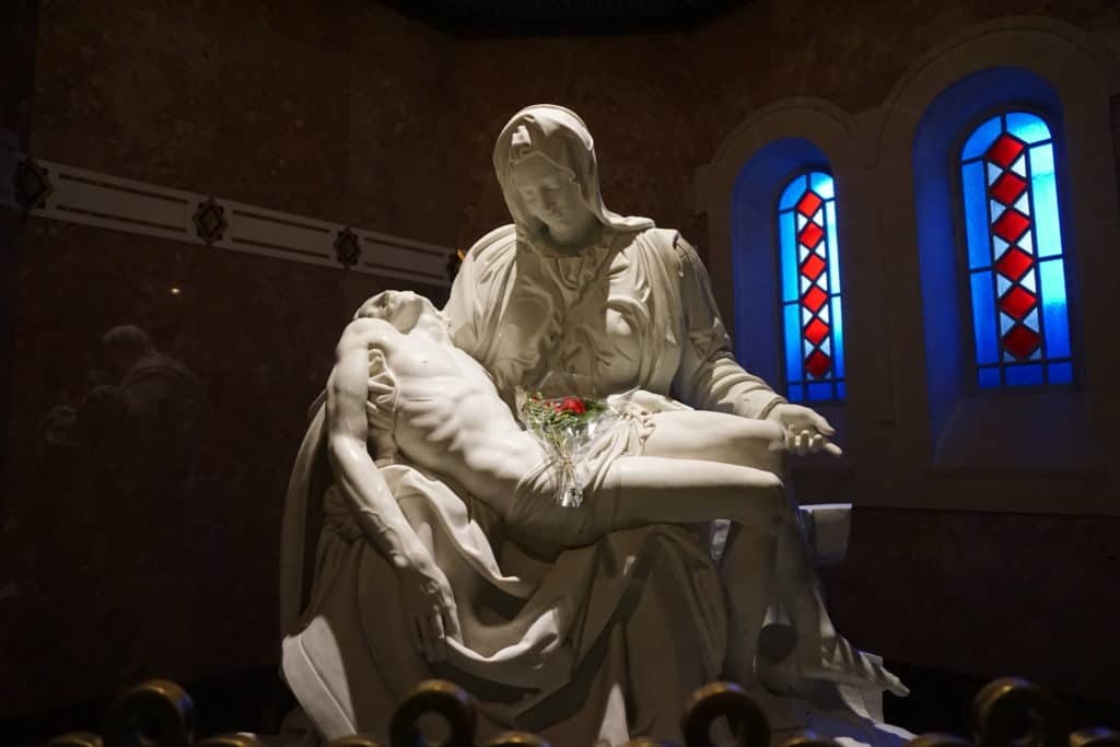 La Pieta sculpture in the Basilica of Sainte-Anne-de Beaupre, Quebec with stained glass windows in background and a bundle of red flowers laying on the body of Jesus.