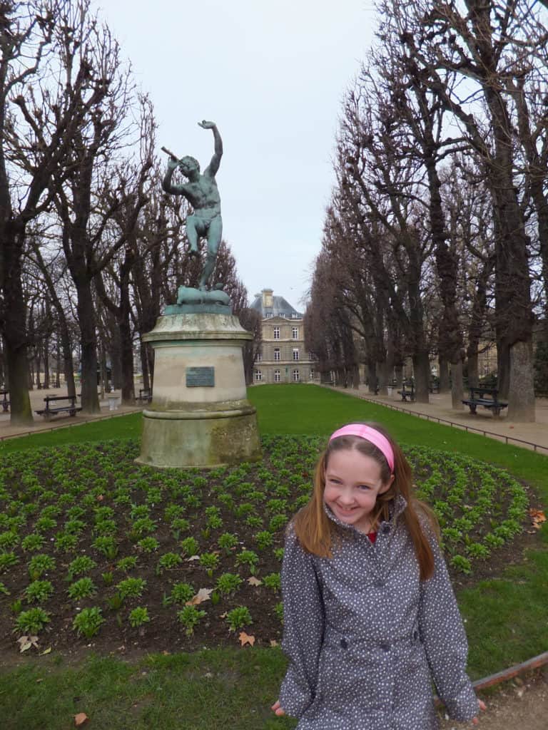 Young girl in rain coat and pink headband standing in Luxembourg Gardens in Paris with statue of Pan behind her.