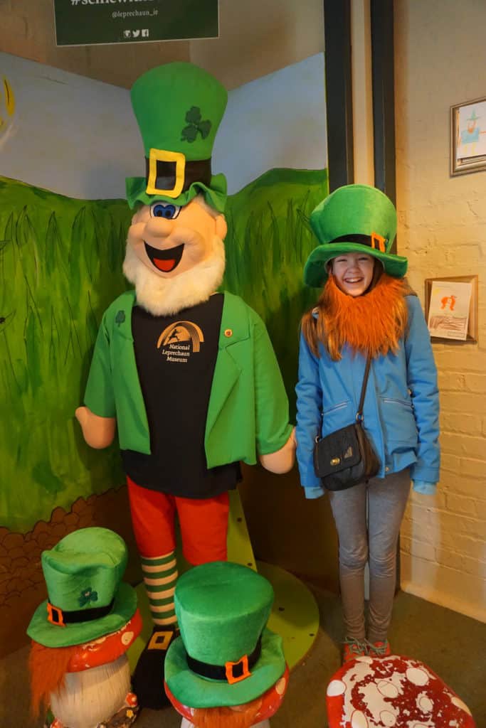 Young girl wearing green hat and red beard standing beside large stuffed toy Leprechaun.