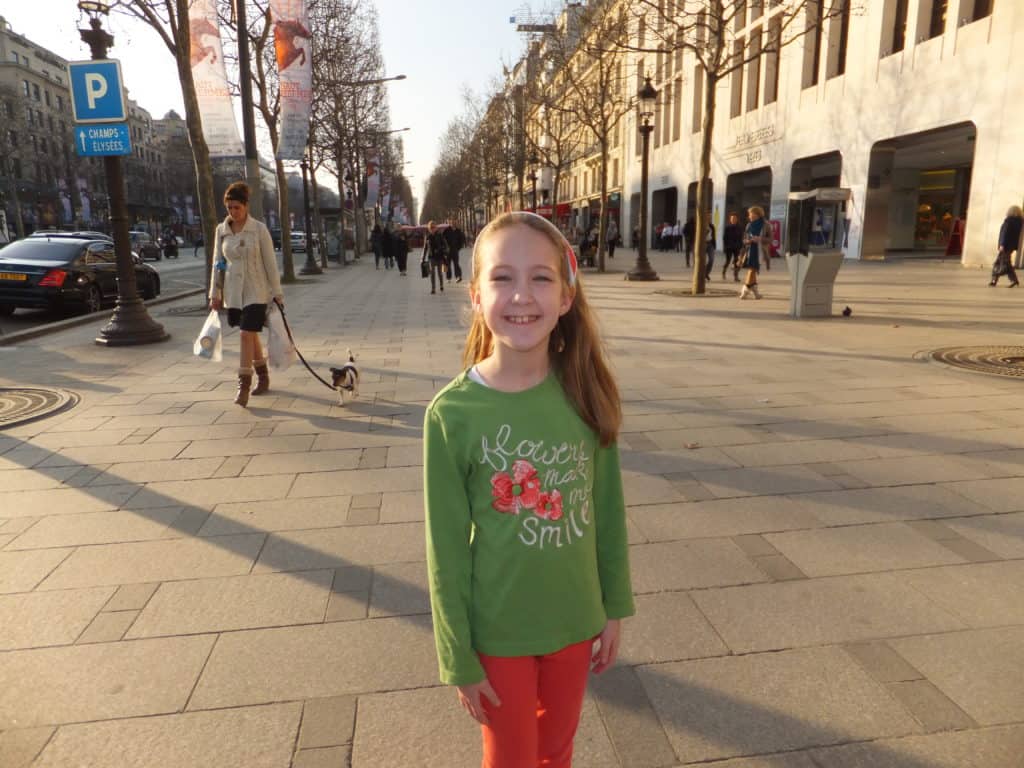 Young girl in green shirt posing on the Champs-Elysee in Paris with woman walking dog on leash in background.