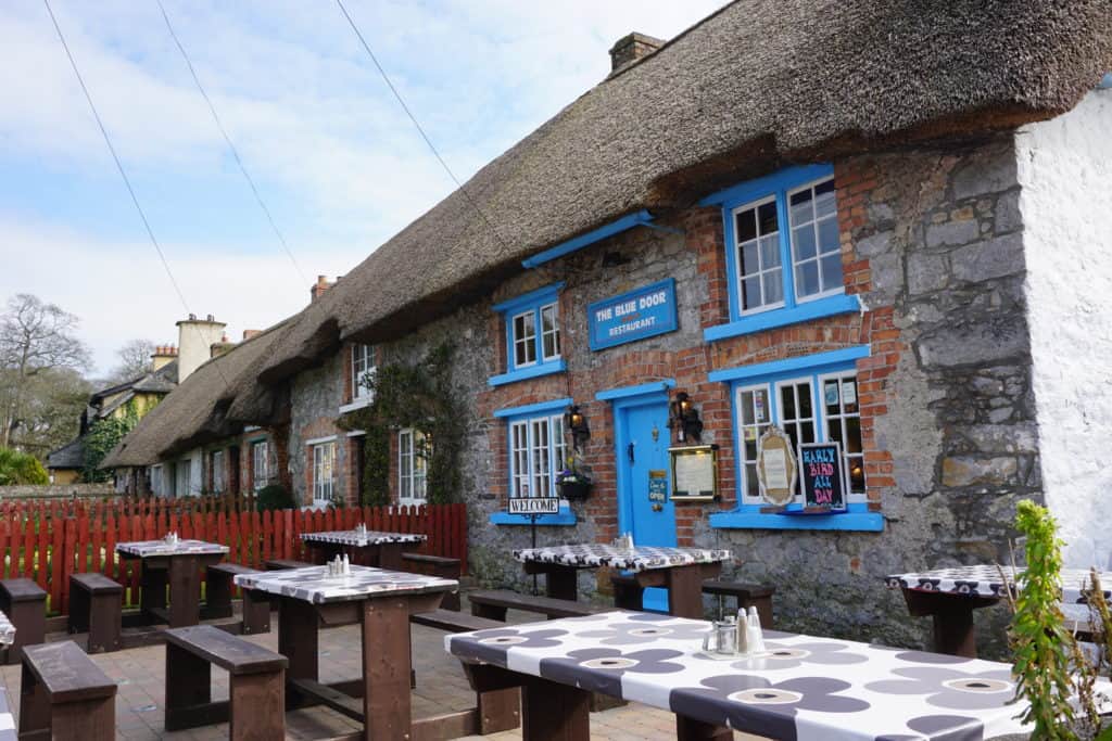 Thatched cottage with blue door and window frame in Adare, Ireland with picnic tables in front garden.