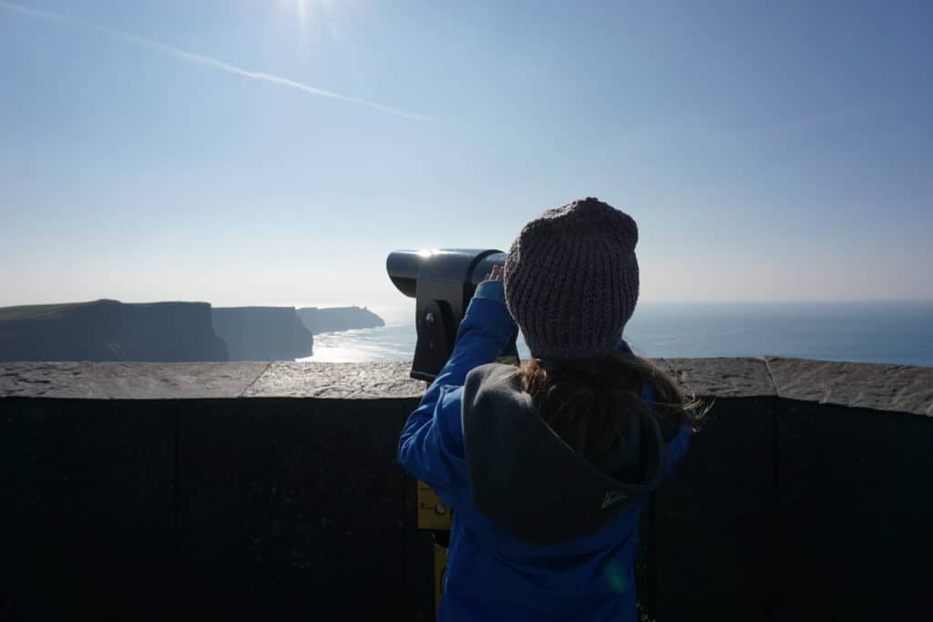 Young girl looking through viewfinder at Cliffs of Moher on a bright sunny day.