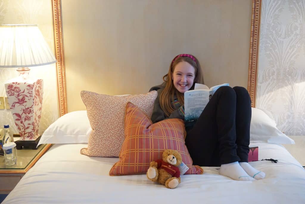 Young girl with book sitting on bed with white bedspread, pink striped and flowered cushions and teddy bear.