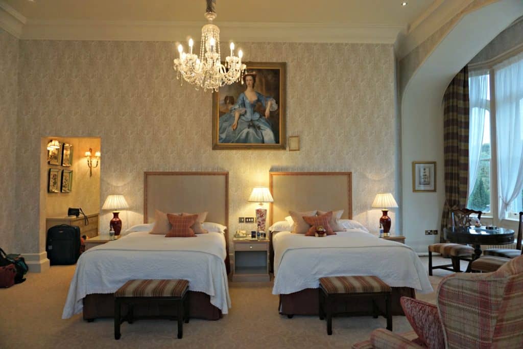 Room at Dromoland Castle - two double beds with white bedspreads and pink cushions in wallpapered room with white chandelier and painting of woman in blue ballgown above beds.