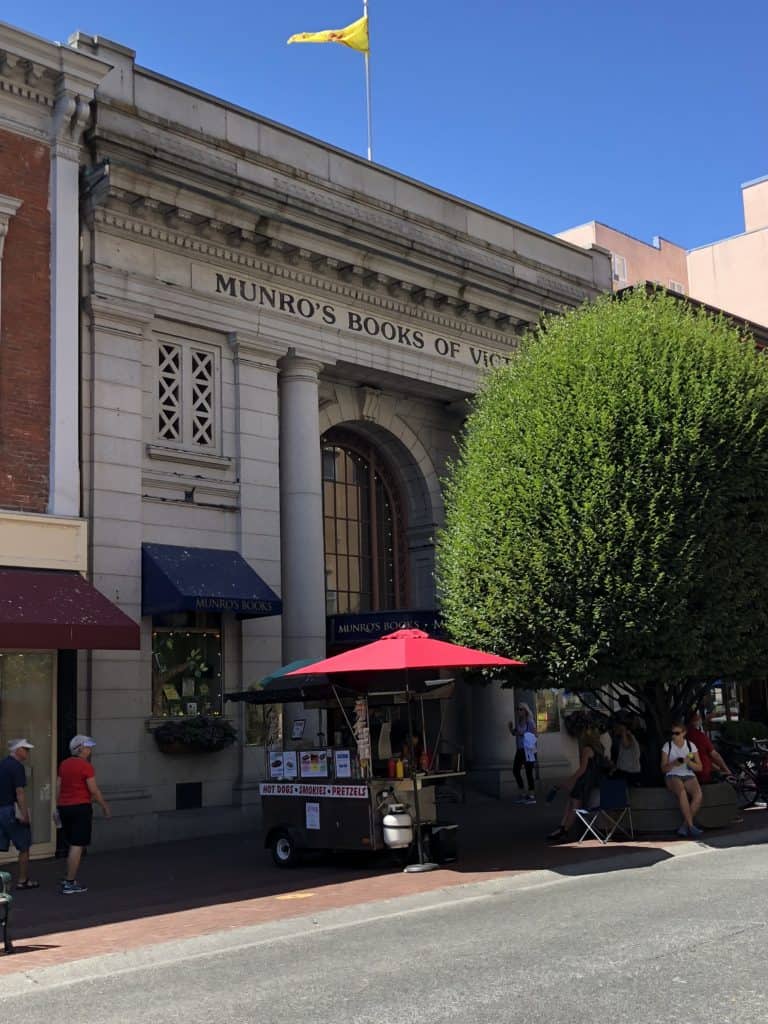 Exterior of Munro's Books of Victoria shop with large tree in front and hot dog cart with red umbrella.