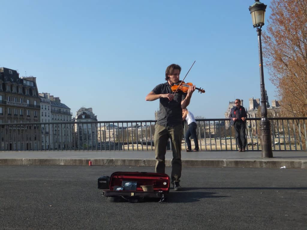 Man playing violin on bridge in Paris with case on ground at his feet and Seine River and buildings in background.