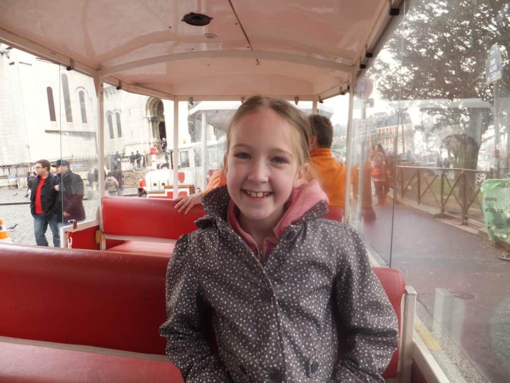 Young girl in rain coat sitting on red bench of the Montmartre tourist train in front of Sacre Coeur Basilica.