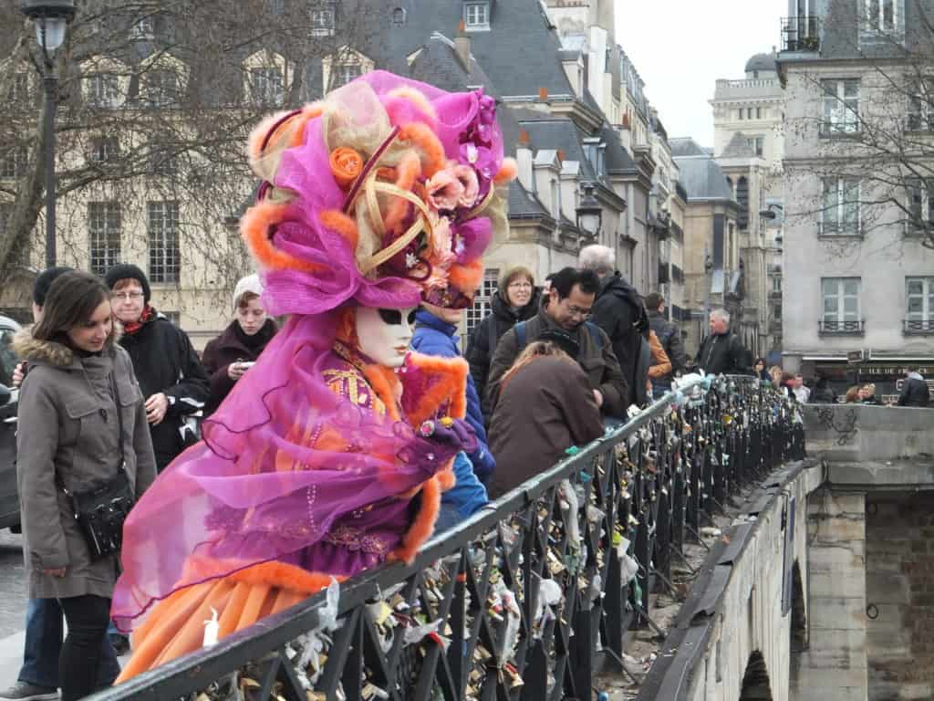 Group of people including woman in a pink and orange carnival costume and white mask looking at love locks on a bridge in Paris.