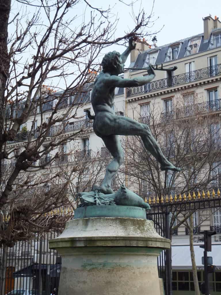 Statue of Pan in Luxembourg Gardens in Paris alongside trees without leaves and Parisian buildings in background.