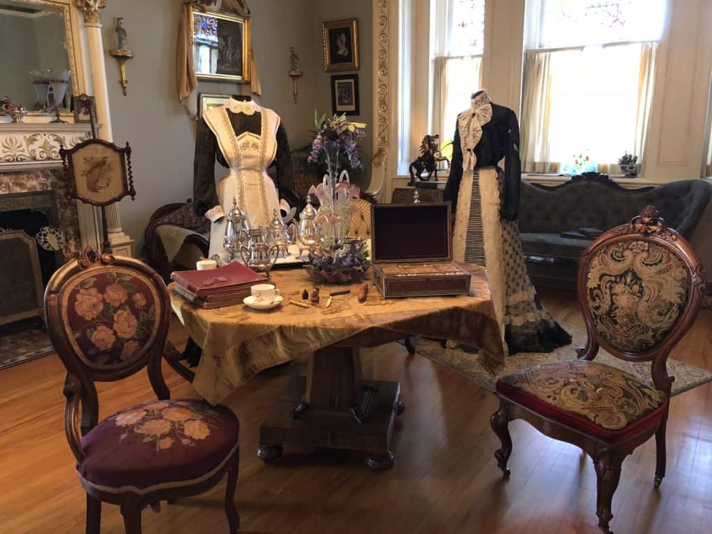 Room display at Craigdarroch Castle - books, tea set on table, needlepoint chairs, two dresses on mannequins.