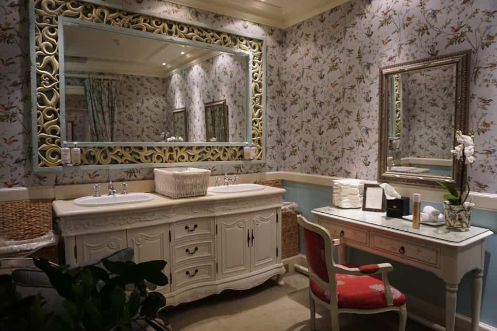 Elaborate powerder room with flowered wallpaper, white cabinet with two sinks and large mirrow and smaller white makeup table with mirror and red upholstered chair.