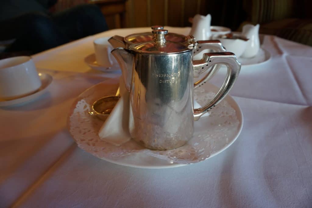 Silver Dromoland Castle teapot sitting on saucer on white tablecloth - white cups, cream and sugar in background.