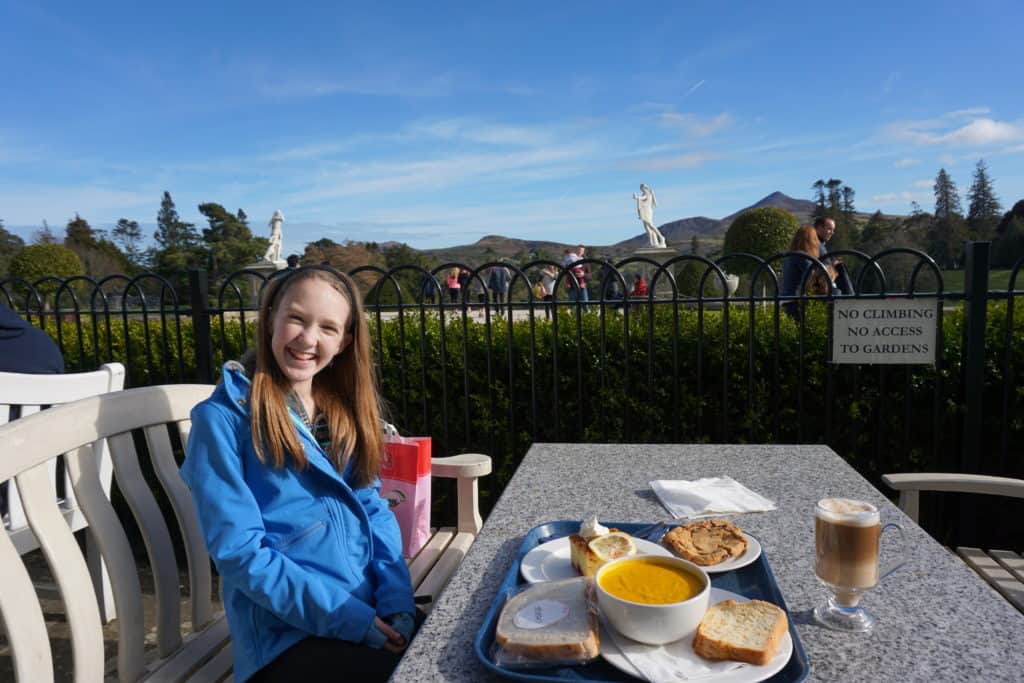 Young girl in blue coat sitting at table in outdoor cafe with tray of food in front of her and Powercourt Gardens in background.