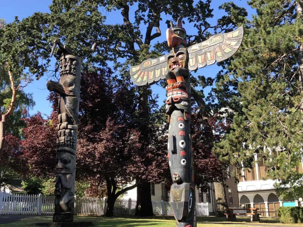 Two totem poles surrounded by trees and buildings in background in Thunderbird Park in Victoria, BC.