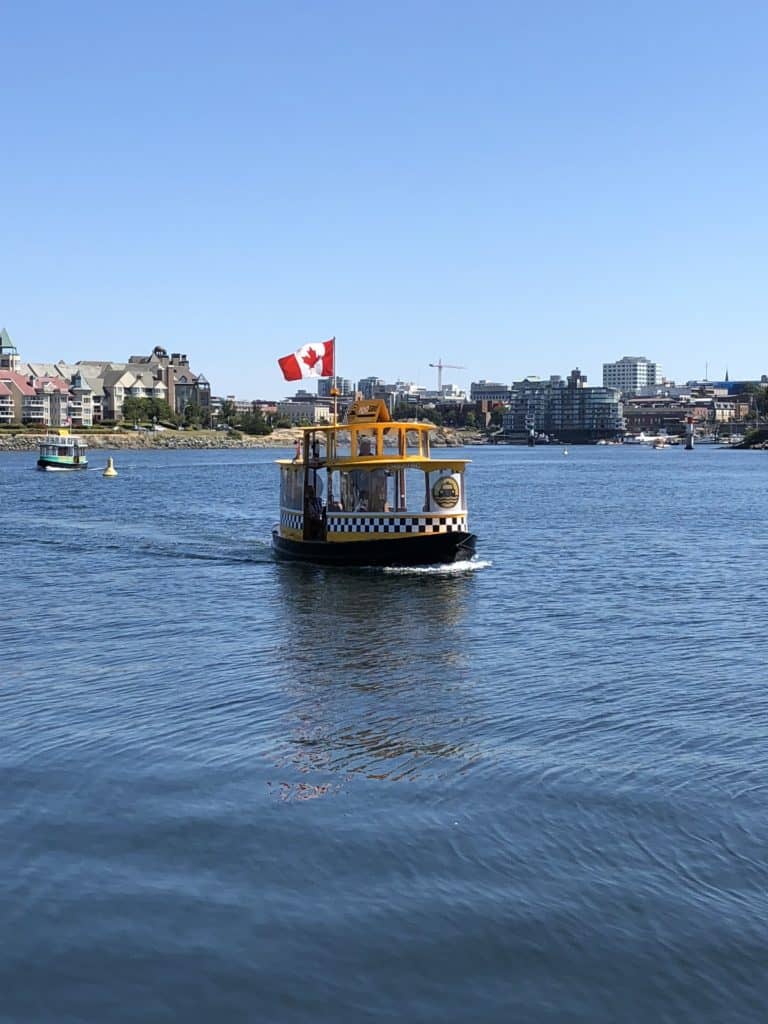 Small water taxi painted like yellow cab flying Canadian flag sailing in Victoria Harbour, Victoria, British Columbia.