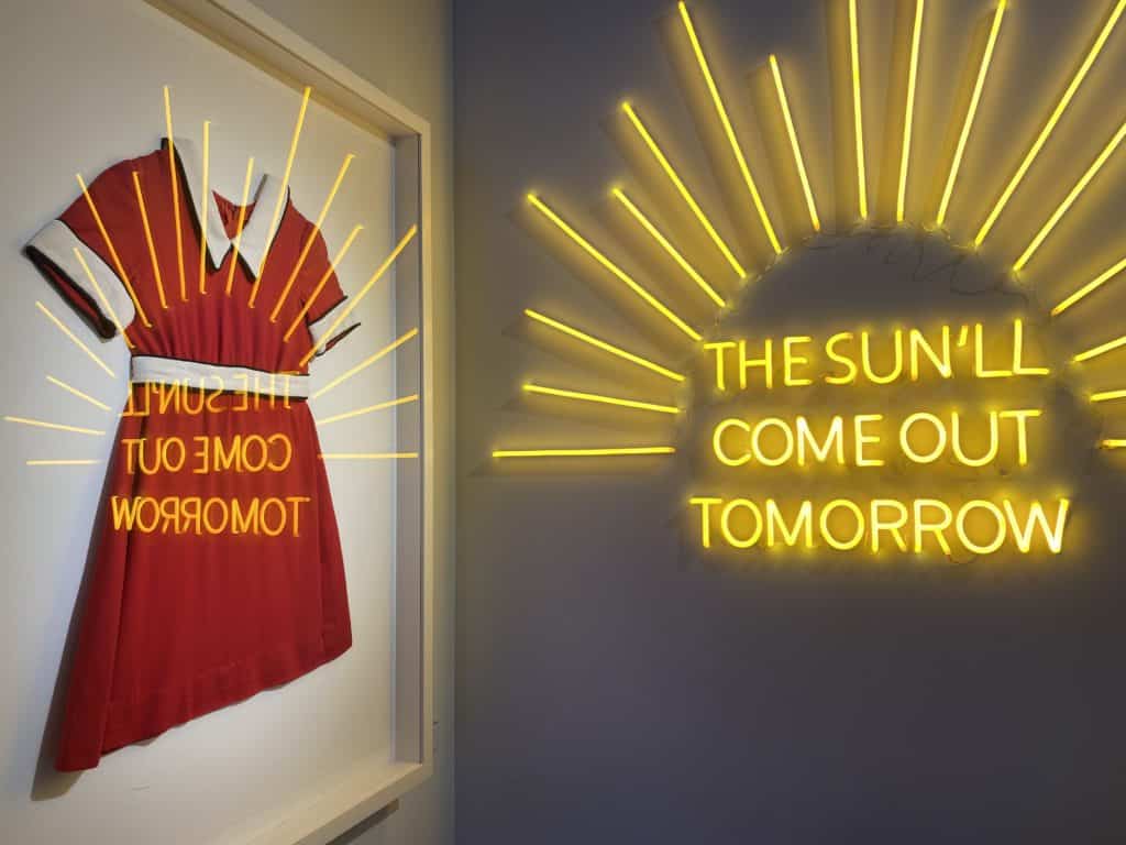 Annie display at the Museum of Broadway - red dress with white trim mounted on wall and yellow lights with rays of sunshine and words reading "The sun'll come out tomorrow" opposite wall and reflected on glass over dress.