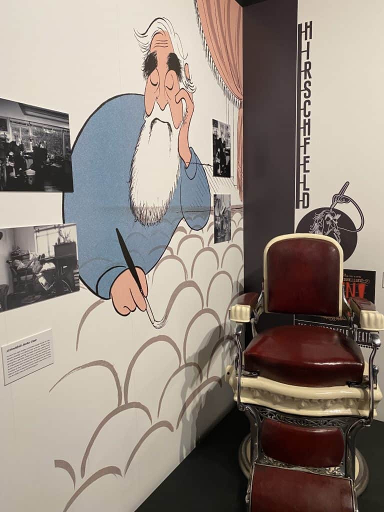 Hirschfeld special exhibit at the Museum of Broadway in New York City - Hirschfeld-style drawing of Hirschfeld painted on wall, display of photos and red chair.