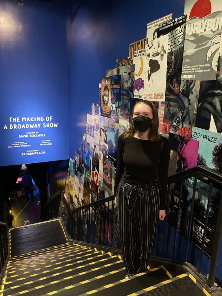 Young woman standing on stairs leading down to The Making of a Broadway Show - walls covered with posters.