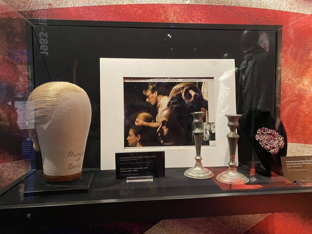 Display case with a blond wig on a stand, photo of wig being placed on Patti Lupone's head, set of candlesticks - Evita the Musical.