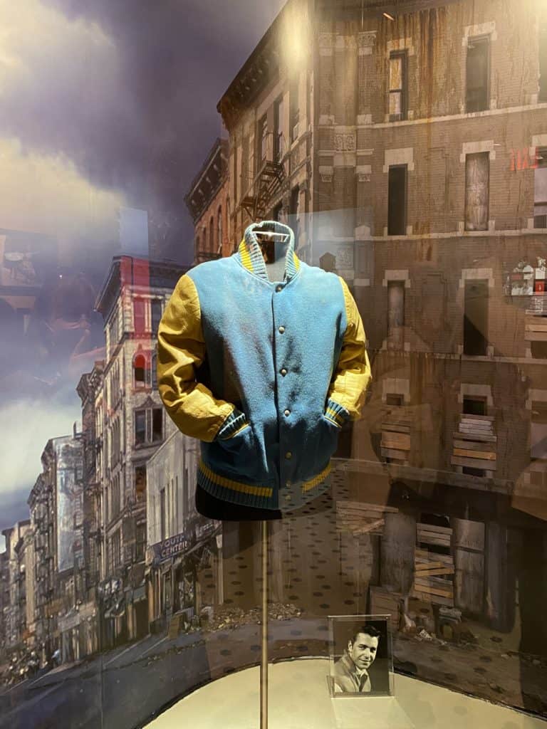 Jacket worn in West Side Story on display with images of New York buildings in background.