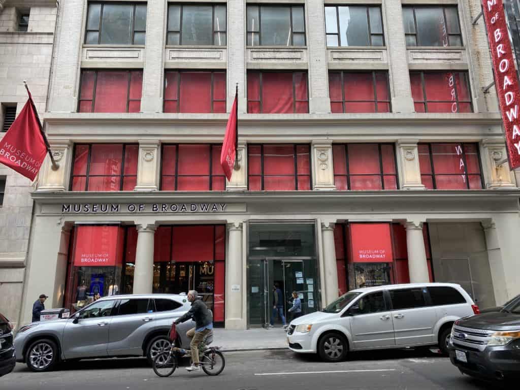 Exterior of the Museum of Broadway in New York City with cars parked in front - red flags, red Broadway sign and windows covered in red.