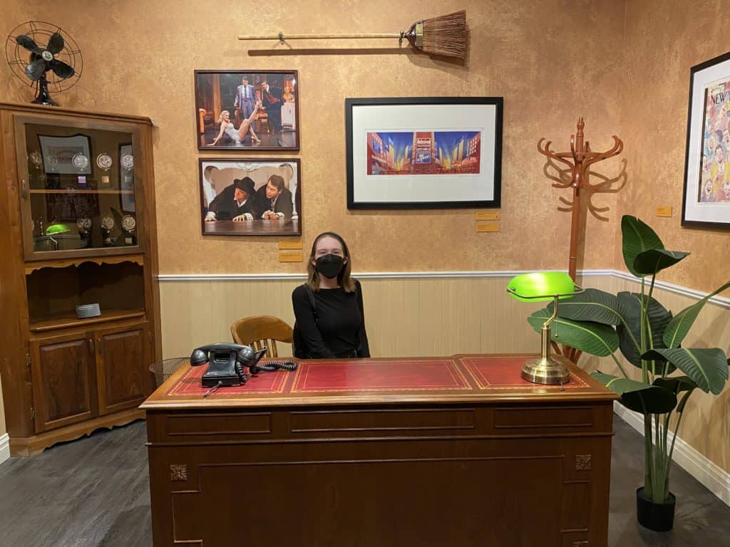 Young woman sitting at desk on The Producers set, Museum of Broadway.