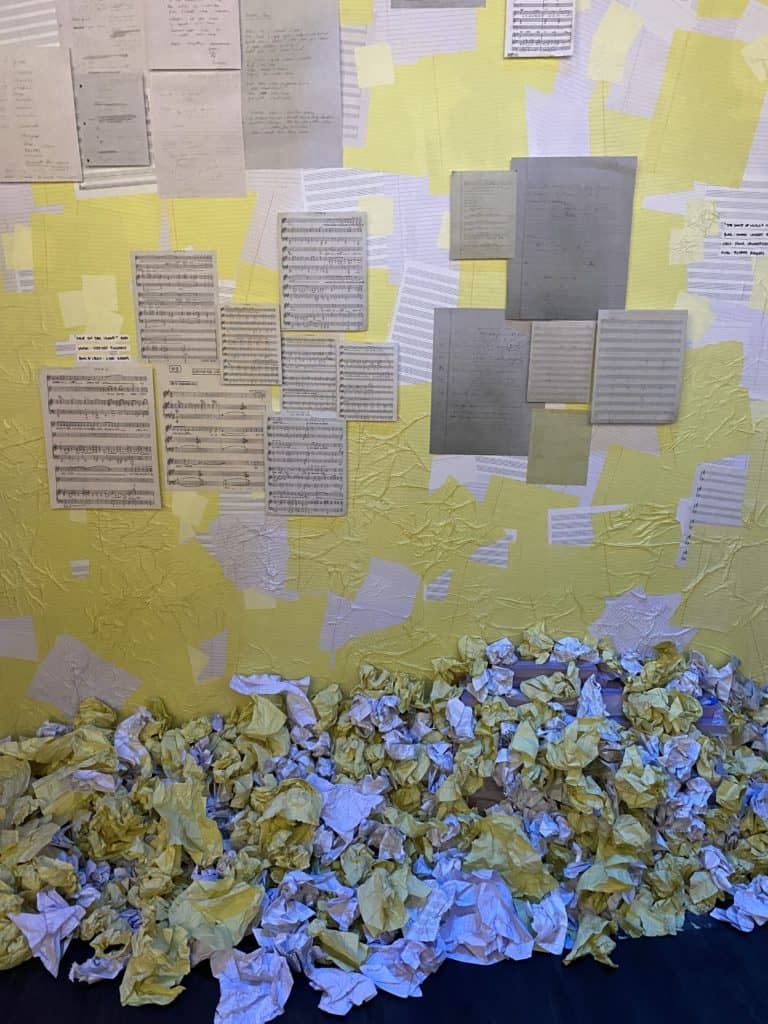 Display of sheet music taped to yellow and white wall with crumpled yellow and white papers on floor.