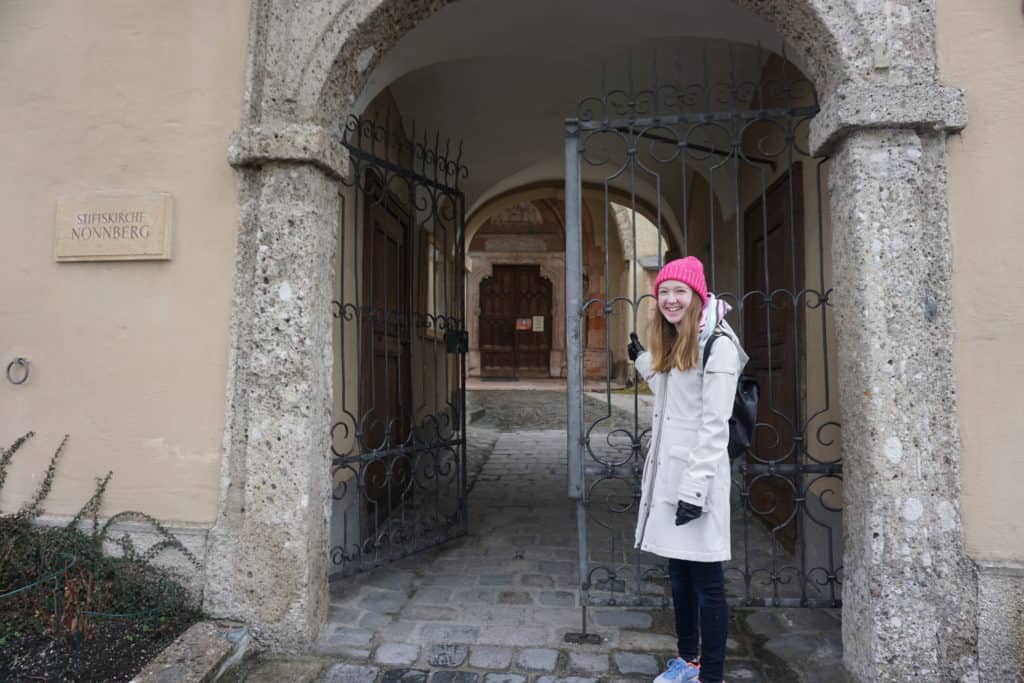 Teen girl in beige coat and bright pink hat opening gate to Nonnberg Abbey in Salzburg, Austria.