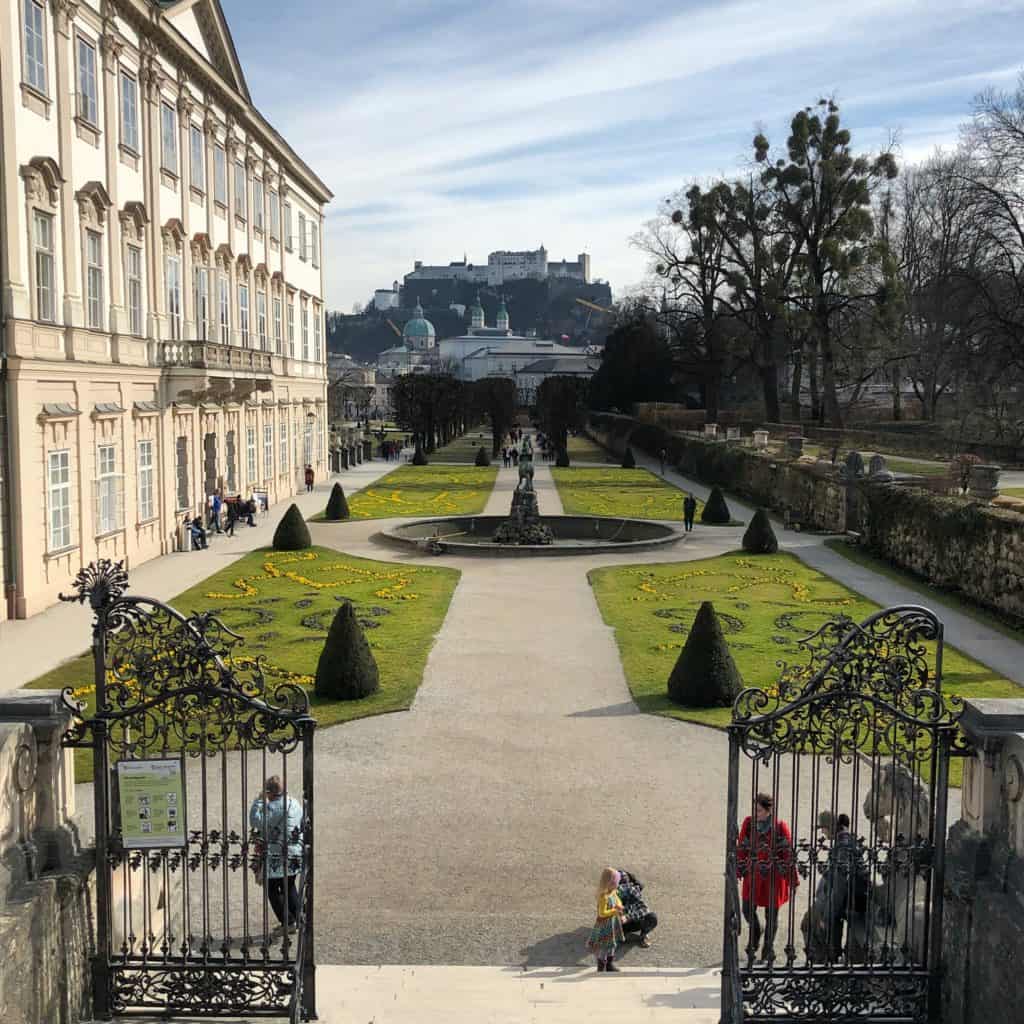 Looking at Mirabell Gardens and Palace from top of the Sound of Music do-re-mi steps with several people standing by iron gates at bottom of steps.