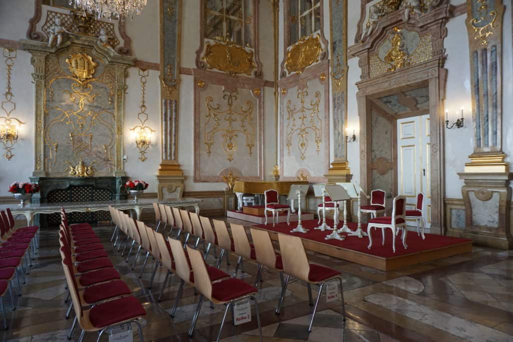 Elaborately decorated room at Mirabell Palace set up with chairs for concert with small stage with chairs and music stands.
