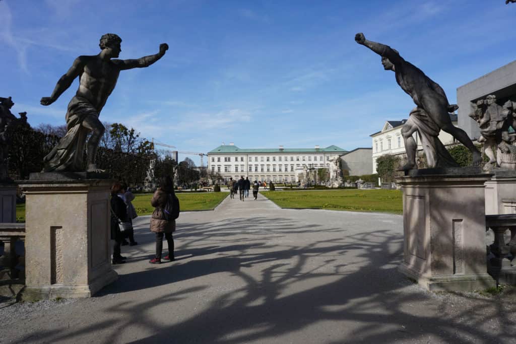 Statues at entrance to Mirabell Gardens in Salzburg, Austria with people walking along paths and Mirabell Palace in background.